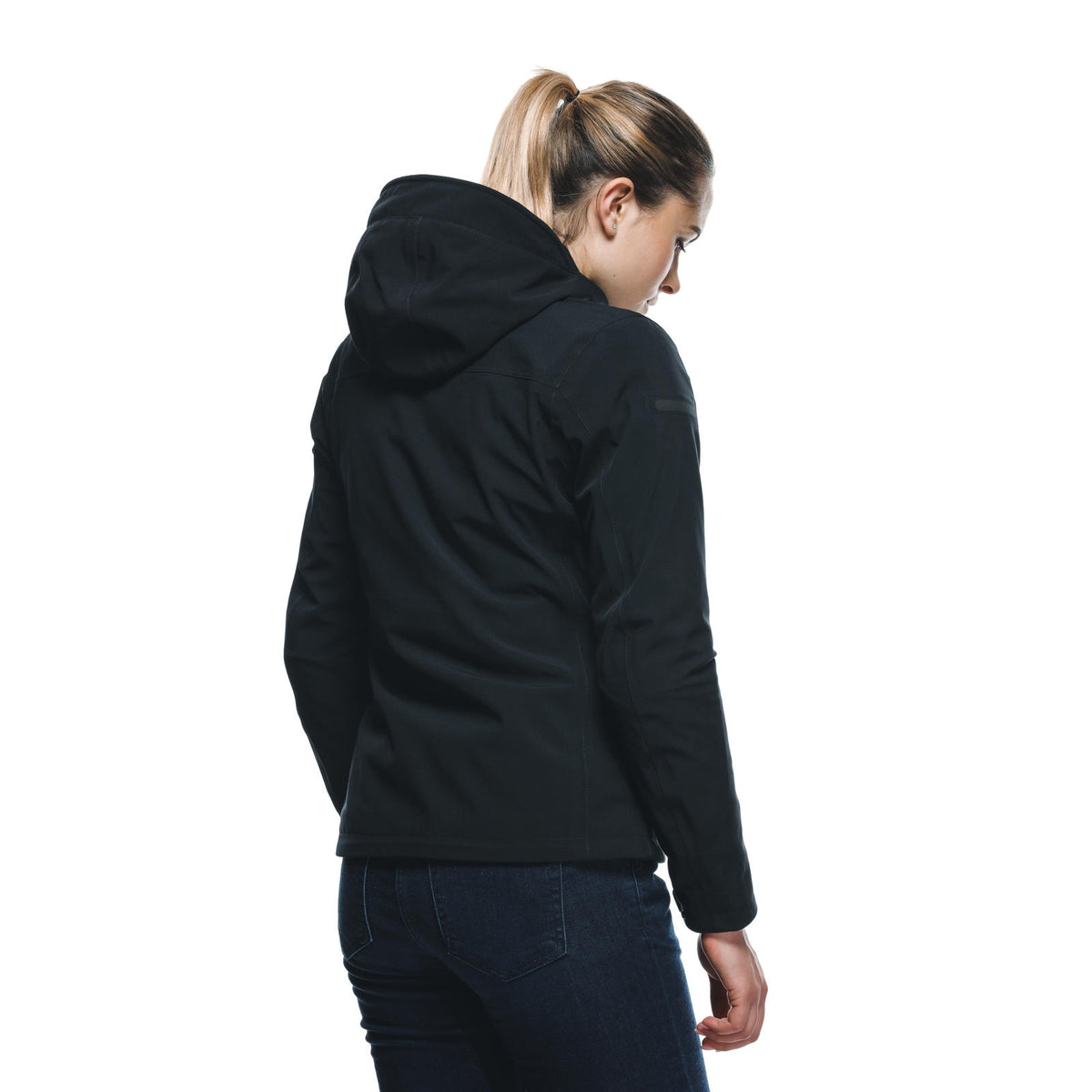 DAINESE CENTRALE ABSØLUTESHELL PRO JACKET WMN 黑色女模特兒背面照,連帽拉起展示背部式樣與臂部反光標籤,可見背心拼接線與肩部護具輪廓,黑色防水布料質感搭配牛仔褲