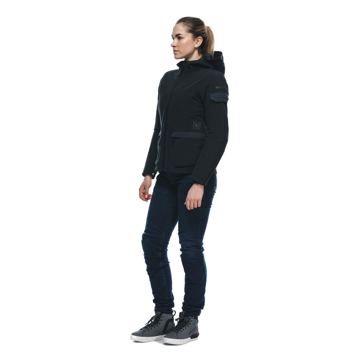 DAINESE CENTRALE ABSØLUTESHELL PRO JACKET WMN 黑色女模特兒側身站姿全身照，連帽拉起展示修身短版外套剪裁，可見臂部口袋與腰間拉鍊設計，搭配牛仔褲與高帮靴，黑色防水布料質感
