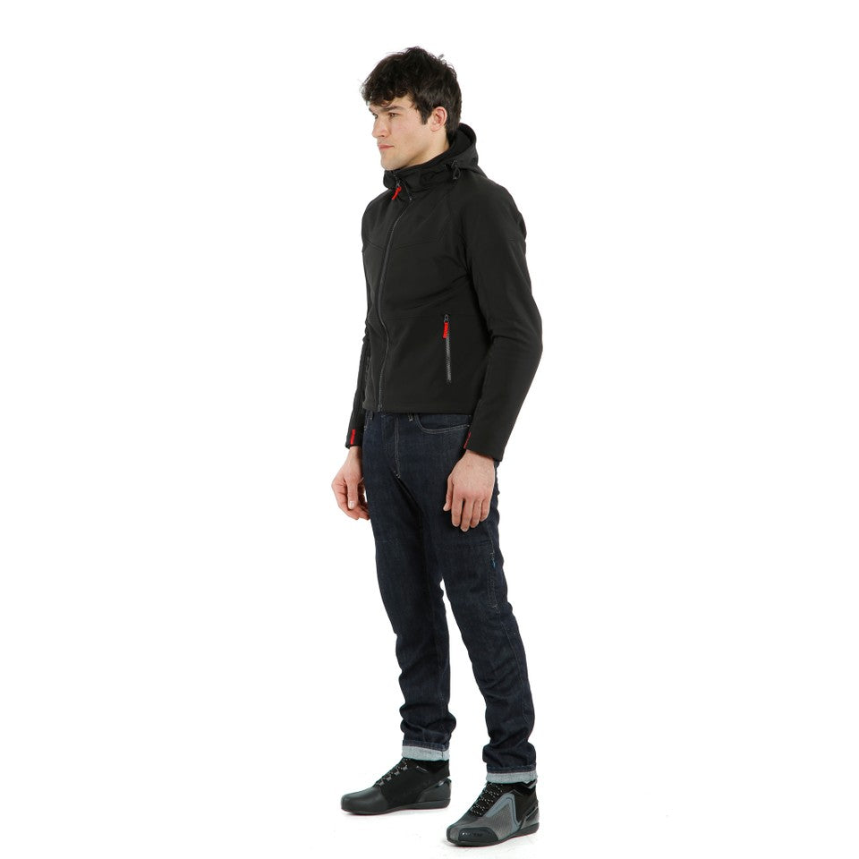 DAINESE IGNITE TEX JACKET 黑/黑配色側面照，男性模特兒側身站立展示外套版型，可見胸前口袋紅色拉鍊拉片與腰際口袋設計，內搭深色牛仔褲與車靴，義大利原廠防風軟殼布料製都會防摔外套，適合日常通勤